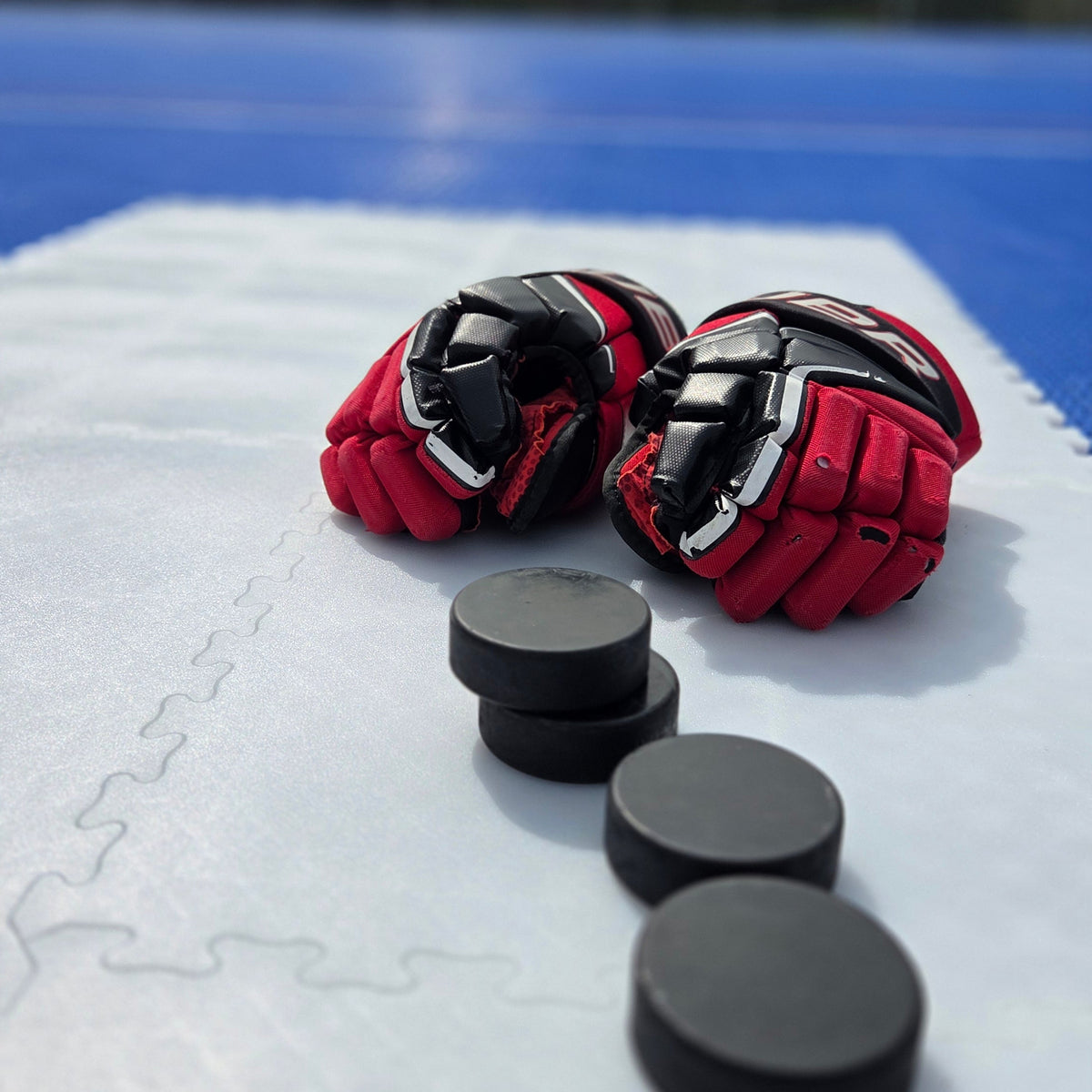Red hockey gloves and black pucks sitting on white interlocking Everyday Ice dryland shooting tiles
